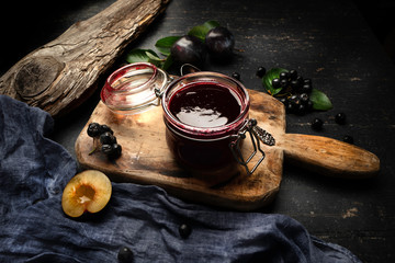 Freshly made plum jam in a jar and garden fruits on a table with drapery. Wood texture.