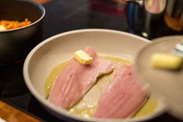 Fisch Filets in Pfanne auf Olivenöl, Butterwürfel darauf, Topf mit Möhren im Hintergrund, Nahaufnahme