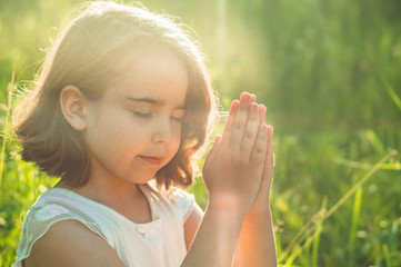 Little Girl closed her eyes, praying in a field during beautiful sunset. Hands folded in prayer concept for faith, spirituality and religion. Peace, hope, dreams concept