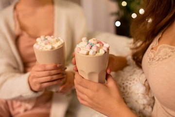 drinks, hygge and pajama party concept - close up of two women or female friends holding cups with hot chocolate with marshmallow at home