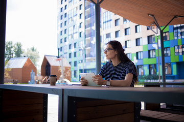 A serious brunette girl, freelancer, with large wrist watch, works at a wooden table on tablet,...