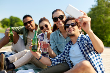 leisure, picnic and technology concept - friends with drinks taking selfie by smartphone on lake pier in summer park