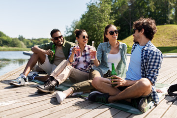 leisure, picnic and people concept - friends drinking beer and cider on lake pier in summer park