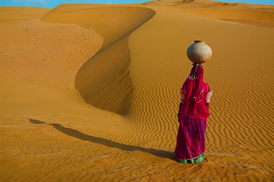 Indian Woman Carrying Heavy Jug Of Water On Her Head And Walking On A Yellow Sand Dune In The Hot Summer Desert Against Blue Sky.water Crises, Jaisalmer, Rajasthan, India