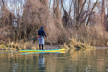 Fototapeta na wymiar Man on stand up paddle boarding (SUP) paddling along the calm autumn Danube river at sunset against a background of trees at the shore. Concept of water tourism, healthy lifestyle and recreation