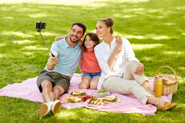 family, leisure and technology concept - happy mother, father and daughter having picnic and taking picture by smartphone on selfie stick at summer park