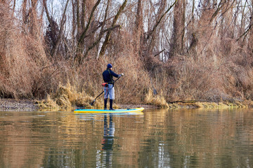 Man on stand up paddle boarding (SUP) paddling along the calm autumn Danube river at sunset against a background of trees at the shore. Concept of water tourism, healthy lifestyle and recreation