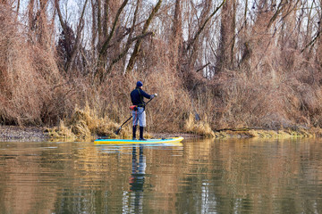 Fototapeta na wymiar Man on stand up paddle boarding (SUP) paddling along the calm autumn Danube river at sunset against a background of trees at the shore. Concept of water tourism, healthy lifestyle and recreation