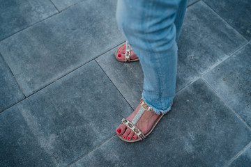 Woman's feet wearing sandals on the paving slabs. Female feet with red pedicure and jeans on legs.