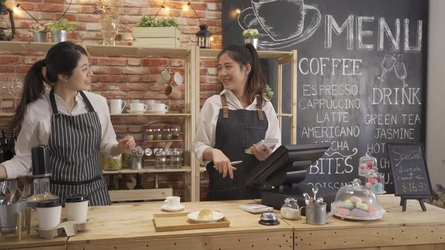two asian women servers in aprons laughing at joke and standing behind counter while working in cozy small bakery together. girl waitress using tablet with customer order and barista prepare coffee