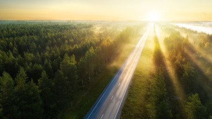 Aerial view of the road in beautiful summer forest at sunrise in Finland. Top view of perfect asphalt roadway and beautiful soft sunlight, green fir trees.