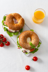 Bagel sandwich with ham, cheese, salad and cherry tomatoes on white chopping board on white background. Bagels and orange juce for breakfast