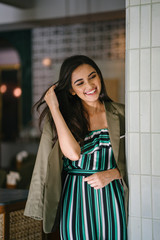 Fototapeta premium Portrait of a tall, slim, elegant and beautiful Indian Asian woman in an elegant jumper with a green jacket draped over her shoulders. She is smiling as she stands in a well-appointed home or room.