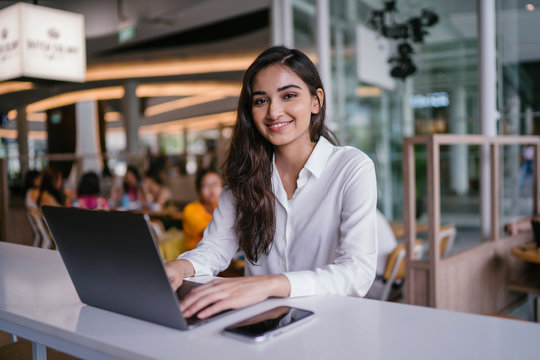 A young and attractive Indian Asian woman small business entrepreneur smiles as she works on her laptop in a cafe during the day. She is wearing a business casual outfit and is hopeful and confident.
