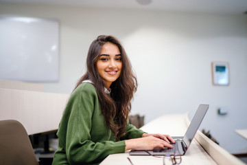 Portrait of a beautiful, young and intelligent-looking Indian Asian woman student wearing a white shirt and green tracker smiling as she works on her laptop in a university classroom. - obrazy, fototapety, plakaty