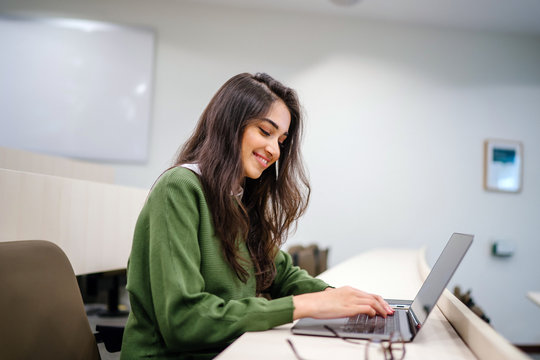 Portrait of a beautiful, young and intelligent-looking Indian Asian woman student wearing a white shirt and green tracker smiling as she works on her laptop in a university classroom.