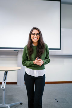Portrait of a young, beautiful, attractive and intelligent Indian Asian woman wearing spectacles in a sweater giving a presentation in a lecture classroom. She is smiling as she is presenting.
