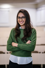 Portrait of a beautiful, young and preppy Indian Asian business woman smiling confidently with her arms crossed in a seminar classroom. She is wearing a preppy outfit and spectacles.