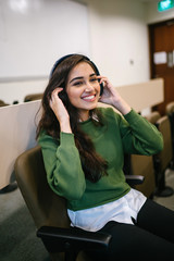 Portrait of a young and attractive Indian Asian woman student in a preppy outfit sitting in a classroom and smiling as she listens to music streaming from her phone to her wireless headphones.