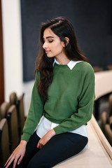 Portrait of a beautiful, young and attractive Indian Asian business woman smiling as she sits in a seminar room during the day. She is wearing a preppy green sweater over a white shirt and jeans.