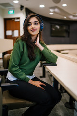 Portrait of a young, beautiful and attractive Indian Asian woman student in a preppy outfit sitting in a room as she listens to music streaming from her phone to her wireless headphones.