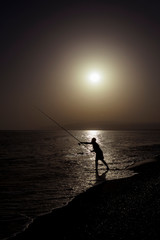 Fototapeta premium Silhouette of fisherman on the beach with fish rod standing on sea water fishing at sunset with beautiful golden sky in vacations relax hobby and leisure holidays concept