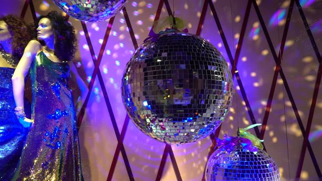 A showcase is decorated with two women mannequins are dressed at a shiny holiday dresses and a disco ball with shiny rays and purple light