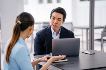 A young, handsome and well-groomed Asian man in a dark suit is interviewing a professional woman candidate applying a job during the day. He is smiling as he talks to the woman.