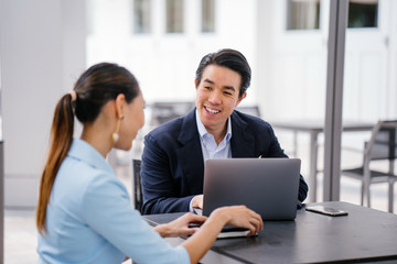 A young, handsome and well-groomed Asian man in a dark suit is interviewing a professional woman candidate applying a job during the day. He is smiling as he talks to the woman.