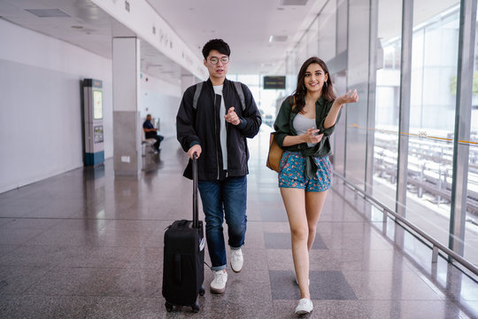A young Asian couple (Korean man, Indian woman) are walking away from the train doors at the airport. They are dressed for a holiday and the man is lugging along his baggage.