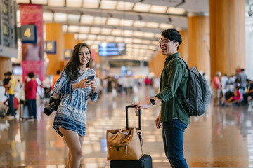 A young interracial diverse couple casually dressed as they stand in the middle of an airport during the day. A young,handsome Korean man and his Indian woman companion are laughing out loud together.