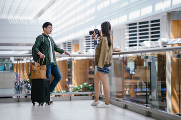 Portrait of a young, diverse and casually dressed interracial Asian couple (a Korean man and Indian woman) standing and taking pictures of one another in a futuristic airport during the day.
