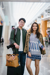 Portrait of a young, attractive and diverse Asian couple (Korean man and his Indian girlfriend) are going for a holiday. They are casually dressed as stand in the middle of an airport during the day.