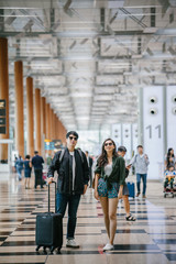 A young and photogenic Asian couple (Korean man, Indian girlfriend in sunglasses) smile as they walk in an airport. They are pulling their luggage behind them on their way for a vacation overseas.