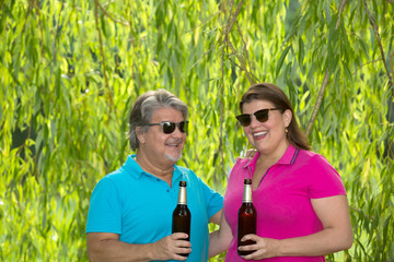 Couple celebrates with bottles of beers while standing in the shade under a tree.