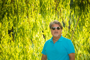 Naklejka na ściany i meble Man poses for a portrait in the shade of a tree. He is serious and looking to the camera. He is wearing sunglasses.