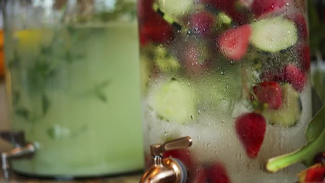 Panning Shot of 3 large glass jars containing different varieties of beverages including strawberry water and lemonade. The liquids are cold as condensation can be seen on the outside of the jars.