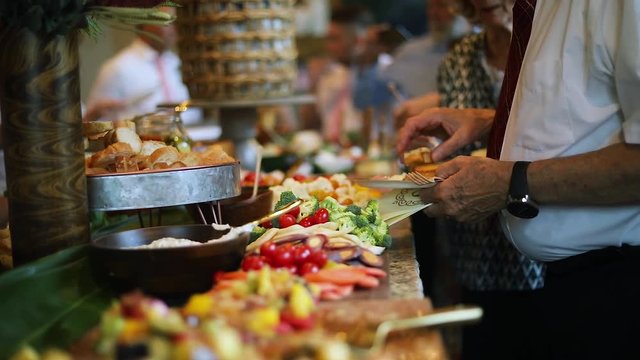 Smooth shot of a line of people at a crowded event going through a food line and grabbing a variety of food including meats, cheeses, bread, fruits, and vegetables.