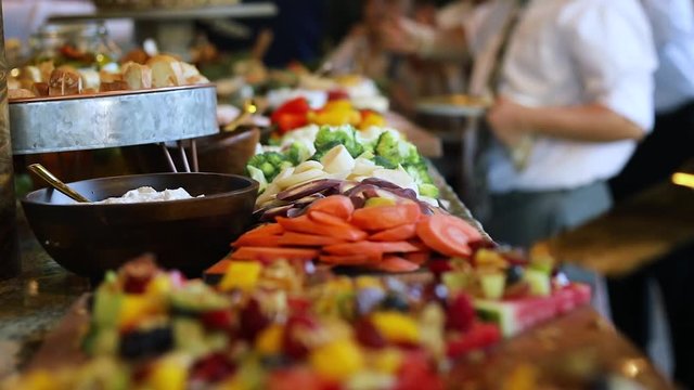 Smooth shot of a line of people at a crowded event going through a food line and grabbing a variety of food including meats, cheeses, bread, fruits, and vegetables.