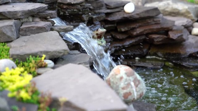 This is a HD slow motion video of a garden waterfall filmed during the Golden Hour in July. It shows a closeup of the water flowing from rocks. There are yellow flowers around the hardscape.