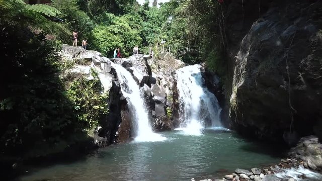 Drone Shot flying towards one of the AlingAling Waterfalls in Bali, Indonesia while someone jumps and does a gainer off of the cliff into the pool of water.