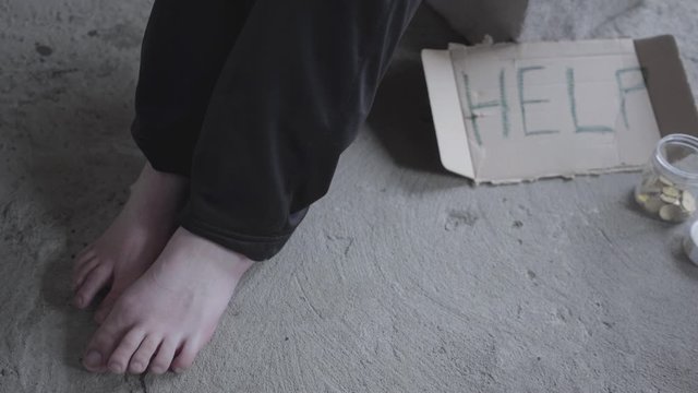 Feet of unrecognized barefoot poor girl sitting in a dusty empty in the abandoned building. A sign that says help and jar with coins lying near. Homeless lady needs help.