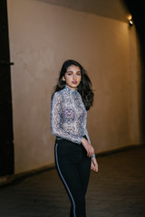 Fashion portrait of a young, beautiful, tall and elegant Indian Asian model in a sheer snakeskin top and black jeans standing and posing against a white wall in the evening. She is smiling softly.