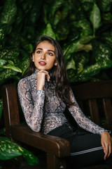 Fashion portrait of a young and beautiful Indian Asian woman in a stylish snakeskin top and black pants sitting on a bench in a park at night. She looks elegant as the flash lights the scene.