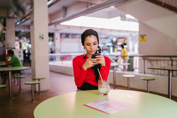 Portrait of a beautiful and attractive Malay Muslim woman in a hijab head scarf sitting and talking on her smartphone as she sips a drink in a hawker center during the day.