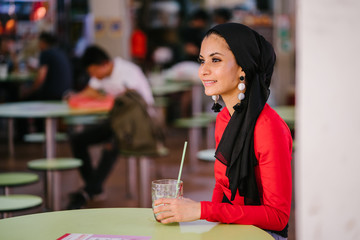 A young and beautiful Middle Eastern Muslim woman wearing a hijab headscarf smiles as she sips a cold drink from a cup as she sits in a hawker center during the day. She is waiting for a friend.