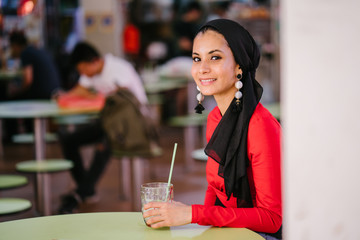A young and beautiful Middle Eastern Muslim woman wearing a hijab headscarf smiles as she sips a...