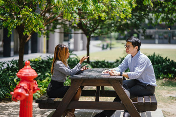 Two colleagues sit and have a business discussion in a park in the city during the day. They are both in office attire. They are a diverse pair -- one a Malay woman, the other a Korean man.