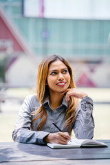 Portrait of a tanned and professional young Southeast Asian business woman in a collared shirt sitting on a bench in the city. She is smiling as she writes and jots in her notebook.