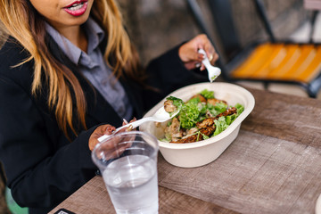 Obraz na płótnie Canvas Close up of a young and a attractive Asian woman in a suit eating a healthy salad lunch with a fork and spoon from a takeaway box. She has a glass of water to go with her salad.